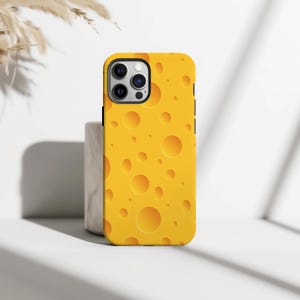 May include: A yellow phone case with a cheese pattern. The case has a bright yellow background with various sized circular holes, mimicking the appearance of Swiss cheese. The phone case is shown on a white surface.