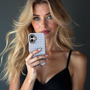 May include: A light blue and white gingham pattern phone case. The case has a black border and a camera cutout. The phone case is held in front of a person with blonde hair and a black top.