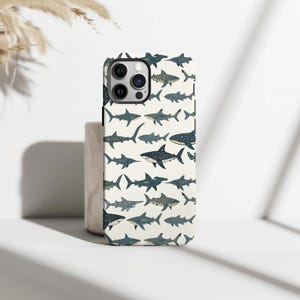 Peut inclure: Coque de smartphone avec un motif répété d'illustrations de requins en nuances de bleu et de gris sur un fond crème. La coque a un bord noir et une découpe pour l'appareil photo.
