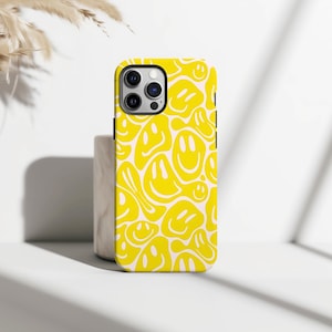 May include: A yellow phone case with a repeating pattern of white smiley faces. The case has a black border and a camera cutout. The background is white with a shadow.