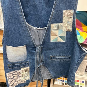 Op de afbeelding: Een blauw spijkerweste met decoratieve patches. De vest heeft een V-hals en een striksluiting. Patches zijn onder meer geometrische en bloemmotieven in blauw, bruin en wit. Voorzien van twee voorzakken en een borstzak.