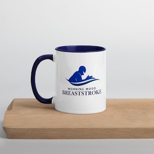 Puede incluir: Taza de cerámica blanca con interior y asa azul marino. La taza presenta un gráfico azul de una persona nadando y las palabras "MORNING MOOD BREASTSTROKE" en una fuente estilizada. La taza está sobre una superficie de madera.