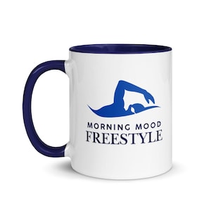 Peut inclure: Mug en céramique blanche avec intérieur et anse bleu marine. La tasse présente une silhouette bleue d'un nageur et les mots "MORNING MOOD FREESTYLE" en bleu marine.
