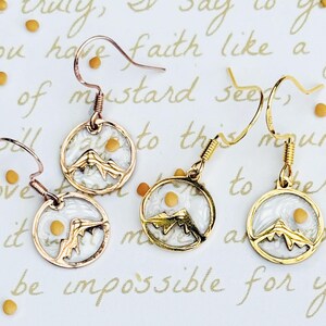 May include: Four pairs of mountain-themed earrings. Two pairs have rose gold hooks and frames, and two have gold hooks and frames. Each earring features a white background with a mountain silhouette and a yellow seed.