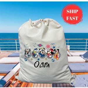 May include: A cream-colored drawstring bag featuring a nautical-themed design with cartoon characters in portholes. The bag has the name "Olivia" printed below the characters. A red circle with the words "SHIP FAST" is visible.