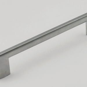 Könnte beinhalten: Ein Schrankgriff aus gebürstetem Nickel. Der rechteckige Griff hat ein gerades Stangendesign mit einem quadratischen Ende auf jeder Seite. Die Hardware hat eine hellsilberne Farbe und ist vor einem weißen Hintergrund platziert.