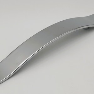 Peut inclure: Une poignée de meuble chromée polie avec un design incurvé en forme de vague. La poignée est rectangulaire avec une surface lisse et réfléchissante. Cette quincaillerie moderne convient aux tiroirs et aux armoires.