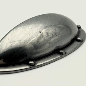Può includere: Una maniglia per armadio in metallo grigio scuro con un design a forma di goccia. La maniglia ha una superficie leggermente strutturata ed è montata su una base abbinata con piccoli rivetti arrotondati. Questo hardware è adatto per il restauro di mobili o nuove costruzioni.