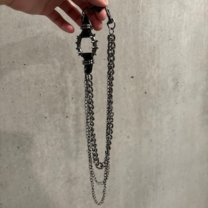 May include: A black wallet chain with a decorative, spiked centerpiece and multiple strands of chain. The chain is held by a hand against a gray background. The metal chain and accents have a dark, gunmetal finish.