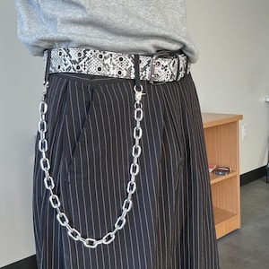May include: A black and white snakeskin print belt with silver grommets and a silver buckle. A silver chain hangs from the belt. The belt is worn with black pinstripe pants.