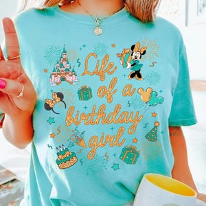 Pu&ograve; includere: Una maglietta azzurra con la scritta dorata "Life of a birthday girl". La maglietta presenta grafiche a cartoni animati di un castello, Minnie Mouse, regali e fuochi d'artificio. &Egrave; visibile una tazza bianca con l'interno giallo.