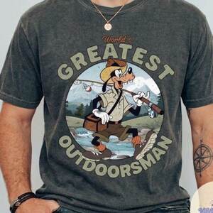 Pu&ograve; includere: T-shirt grigio scuro con un cartone animato di Pippo come pescatore, con la scritta "World's Greatest Outdoorsman" in un design circolare. La grafica include una canna da pesca, una scatola per attrezzi e un paesaggio montano.