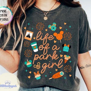 May include: Dark gray t-shirt with the text "life of a park girl" and various cartoon graphics in orange and teal. The shirt also has the words "More Color Options" and "Pepper" on it.