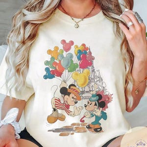 May include: Cream-colored t-shirt featuring a colorful graphic of Mickey and Minnie Mouse. Mickey is holding a bunch of Mickey Mouse-shaped balloons. A castle is in the background. The shirt has a relaxed fit and short sleeves.