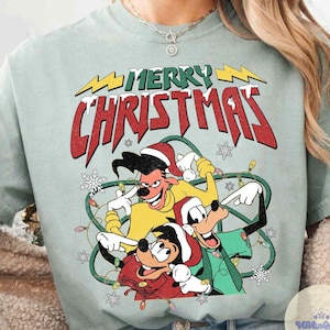 May include: A light green t-shirt featuring a cartoon graphic with the text "MERRY CHRISTMAS". The graphic includes three cartoon characters wearing Santa hats and surrounded by Christmas lights and snowflakes.