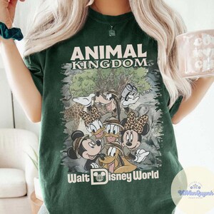 K&ouml;nnte beinhalten: Dunkelgr&uuml;nes T-Shirt mit der Aufschrift "ANIMAL KINGDOM" und "Walt Disney World". Das Shirt hat eine Grafik von Mickey, Minnie, Donald, Daisy, Goofy und Pluto. Die Grafik hat ein Safari-Thema.