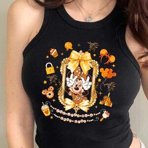 May include: Black tank top featuring a Minnie Mouse graphic in a gold frame. The design includes a bow, fireworks, and other Disney-themed elements. The tank top is a cropped style.