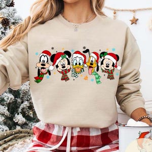 May include: Beige sweatshirt featuring a festive graphic of Disney characters, including Mickey Mouse, Donald Duck, and Pluto, all wearing Santa hats and scarves. The characters are arranged in a row, with snowflakes and stars around them. The sweatshirt is paired with red and white plaid pajama pants.
