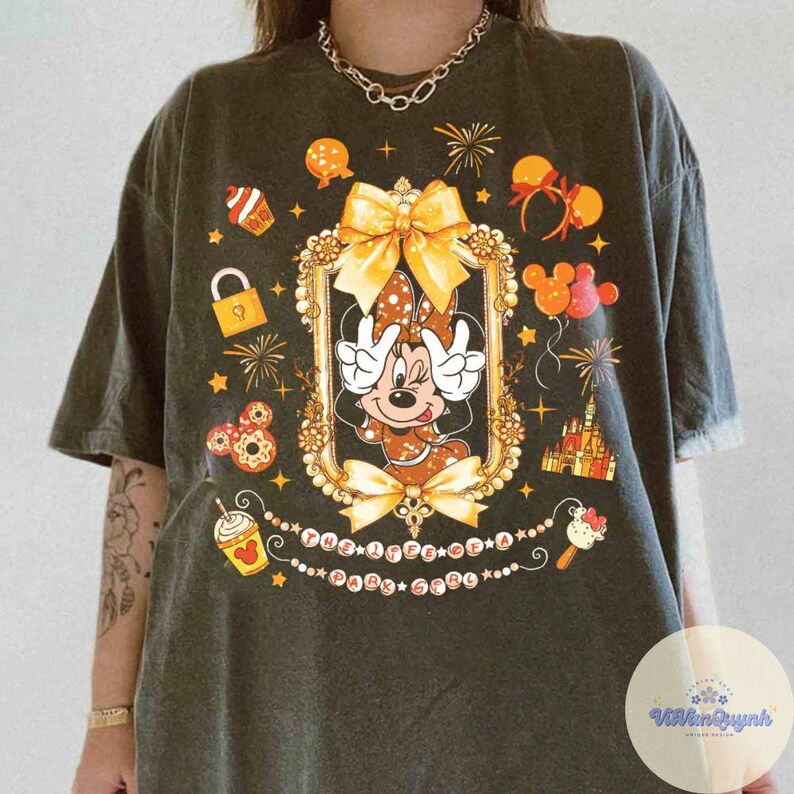 May include: Dark gray t-shirt featuring Minnie Mouse in a gold-framed design with a bow. The shirt includes various Disney-themed graphics like fireworks, cupcakes, and a castle. The text "THE HAPPY BABY GIRL" is also present.