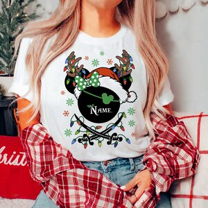 May include: White t-shirt featuring a festive design with a black Mickey Mouse head wearing a Santa hat and reindeer antlers. The design includes a green bow, snowflakes, and string lights. The word "Name" is printed on the design.