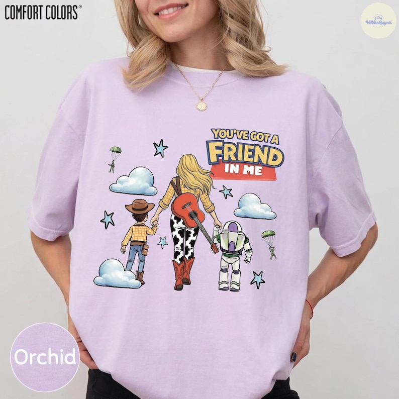 Op de afbeelding: Een lichtpaars t-shirt met een afbeelding van een persoon met lang blond haar die hand in hand loopt met een cowboy en een space ranger. Het ontwerp bevat een gitaar, wolken, sterren, parachutes en de tekst You've Got A Friend In Me.