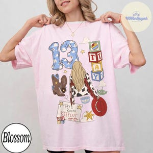 May include: A light pink t-shirt featuring a graphic print of the number 13 in a blue sky pattern, a person with long blonde hair, cowboy boots, alphabet blocks, a green alien, and a sign that reads Tay's Room Keep Out except you.