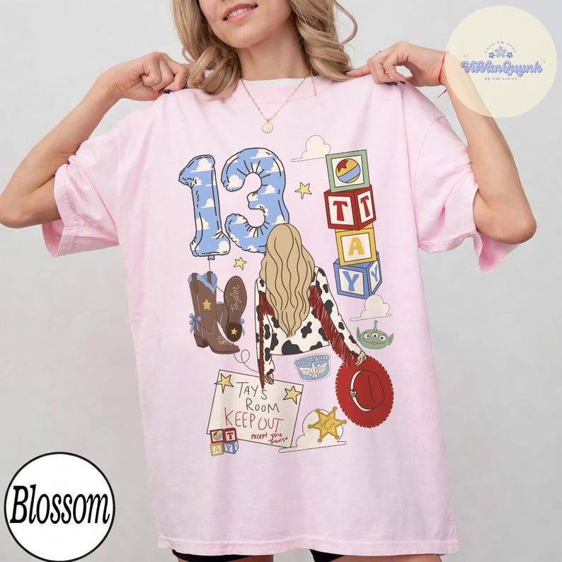 May include: A light pink t-shirt featuring a graphic print of the number 13 in a blue sky pattern, a person with long blonde hair, cowboy boots, alphabet blocks, a green alien, and a sign that reads Tay's Room Keep Out except you.
