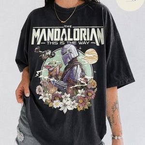 K&ouml;nnte beinhalten: Schwarzes T-Shirt mit einem Aufdruck von The Mandalorian und Grogu, umgeben von Blumen. Der Text "THE MANDALORIAN - THIS IS THE WAY" ist oben aufgedruckt. Das T-Shirt ist schwarz.