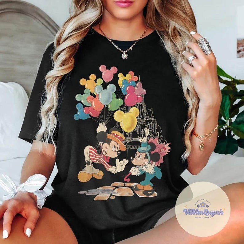 May include: Black t-shirt featuring a colorful graphic of Mickey and Minnie Mouse. Mickey Mouse is holding a bouquet of balloons, and Minnie Mouse is holding a parasol. A castle is in the background.