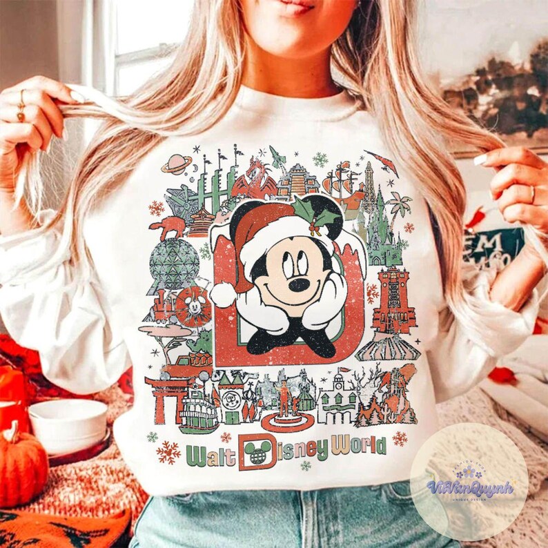 May include: White sweatshirt featuring a festive Mickey Mouse design. Mickey is wearing a Santa hat and surrounded by iconic Walt Disney World landmarks. The words "Walt Disney World" are printed at the bottom in green and red.
