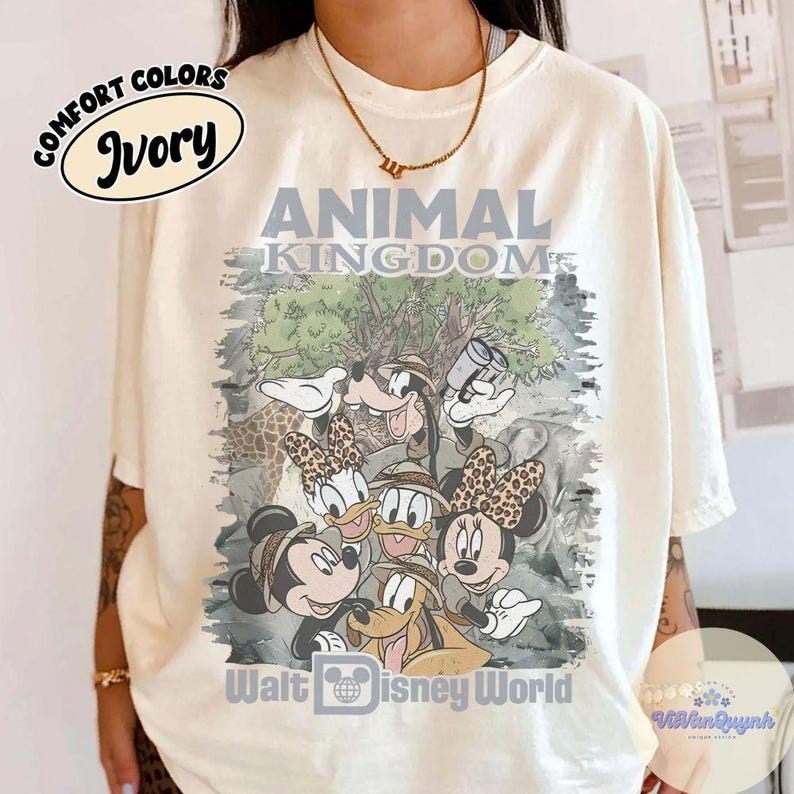K&ouml;nnte beinhalten: Elfenbeinfarbenes T-Shirt mit einer Grafik von Disney-Figuren in Safari-Kleidung, darunter Mickey, Minnie, Donald, Daisy, Goofy und Pluto. Das Shirt tr&auml;gt den Text "ANIMAL KINGDOM" und "Walt Disney World". Das Shirt besteht aus Comfort Colors-Material.