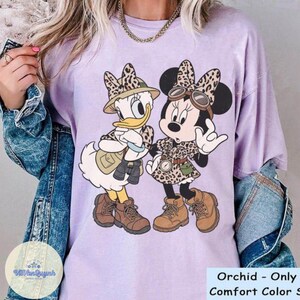 May include: A light purple t-shirt featuring a graphic of Daisy Duck and Minnie Mouse in safari outfits. The characters are wearing safari hats, dresses, and boots. The text "Orchid - Only Comfort Color S" is visible at the bottom.