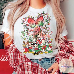 May include: White t-shirt featuring a festive Disney-themed design. The graphic includes Mickey and Minnie Mouse, Donald Duck, and other characters in holiday attire, with a castle and a bow-shaped Christmas light design.