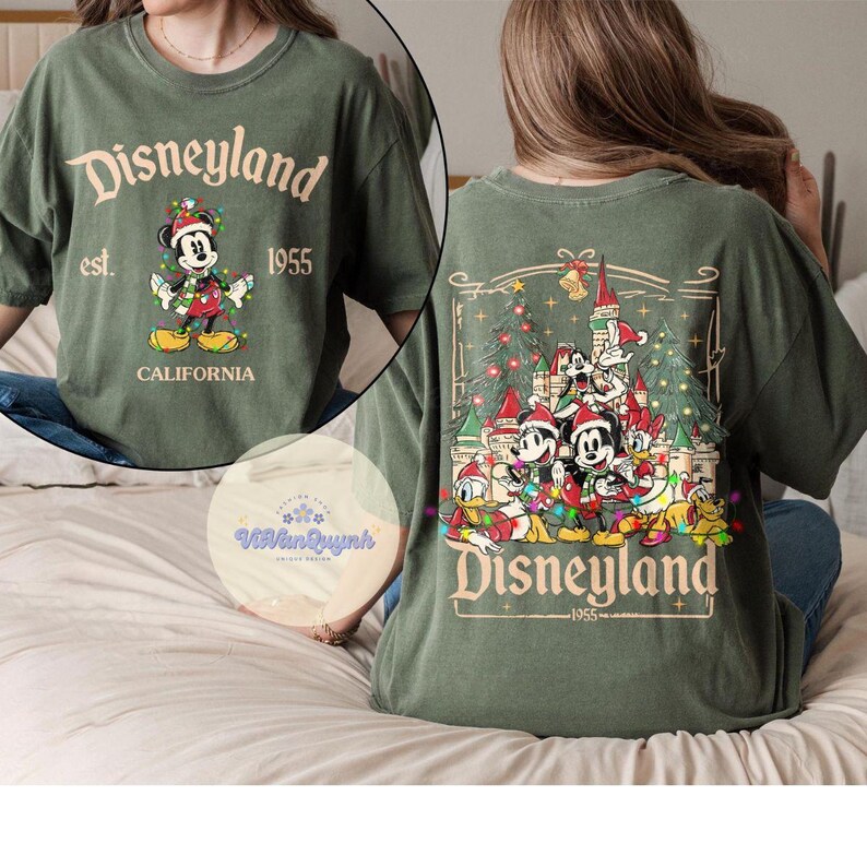 May include: Olive green Disneyland t-shirt featuring Mickey Mouse in a Christmas design. The front of the shirt has the text "Disneyland est. 1955 California". The back of the shirt has a Christmas scene with Disney characters.