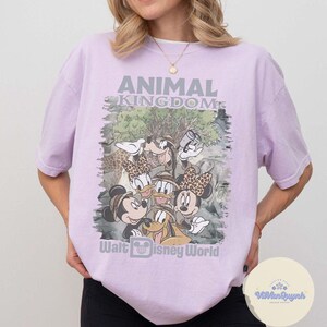K&ouml;nnte beinhalten: Ein helllila T-Shirt mit einer Grafik von Disney-Figuren, darunter Mickey und Minnie Mouse, Donald Duck und Goofy. Der Text "ANIMAL KINGDOM" und "Walt Disney World" ist auf der Vorderseite aufgedruckt.