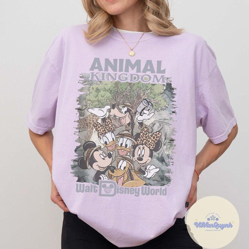 K&ouml;nnte beinhalten: Ein helllila T-Shirt mit einer Grafik von Disney-Figuren, darunter Mickey und Minnie Mouse, Donald Duck und Goofy. Der Text "ANIMAL KINGDOM" und "Walt Disney World" ist auf der Vorderseite aufgedruckt.