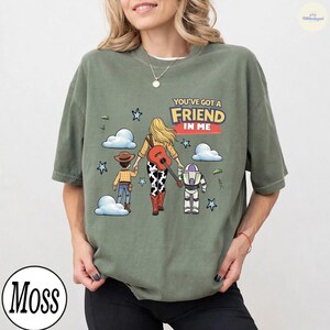 Op de afbeelding: Een olijfgroen T-shirt met ronde hals en een grafische illustratie van een blond persoon die hand in hand loopt met een cowboyfiguur en een ruimtevaarderfiguur. De tekst "YOU'VE GOT A FRIEND IN ME" staat boven de figuren met blauwe sterren.
