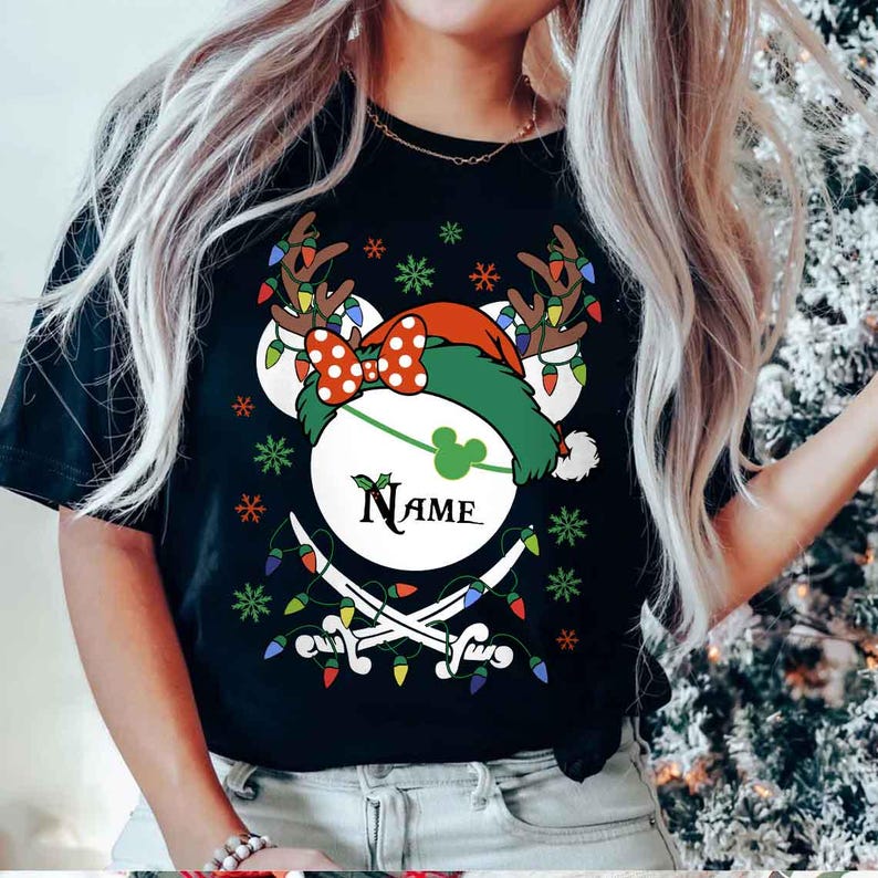 May include: Black t-shirt featuring a festive design. The design includes a white circle with a green Mickey Mouse silhouette, a red and white polka dot bow, and a green pirate hat. The word "NAME" is written in a script font. The design also includes crossed swords and Christmas lights.