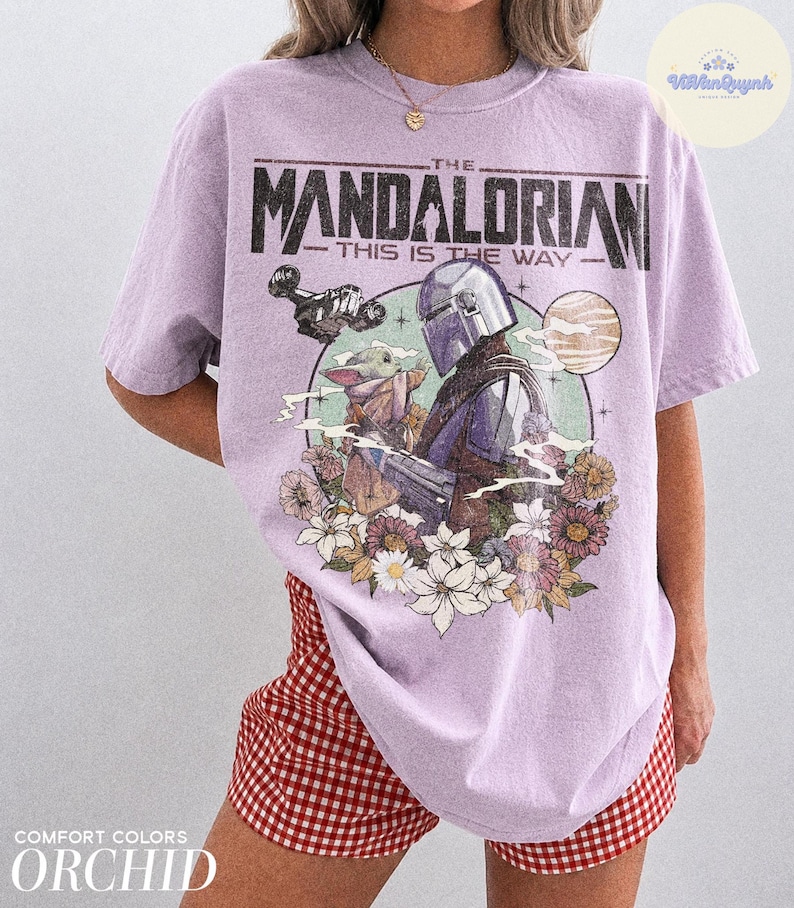 K&ouml;nnte beinhalten: Orchideenfarbenes T-Shirt mit einem Grafikdesign aus "The Mandalorian". Das Design zeigt den Mandalorianer, Grogu und florale Elemente. Der Text "THE MANDALORIAN - THIS IS THE WAY" ist oben aufgedruckt. Das Shirt ist von der Marke Comfort Colors.