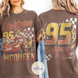 K&ouml;nnte beinhalten: Braunes T-Shirt mit einem Used-Look-Grafik von Lightning McQueen aus dem Film Cars. Das Design umfasst die Nummer 95, den Namen "Lightning McQueen" und eine Zielflagge. Auf der R&uuml;ckseite des Shirts ist ebenfalls die Nummer 95 und der Name zu sehen.