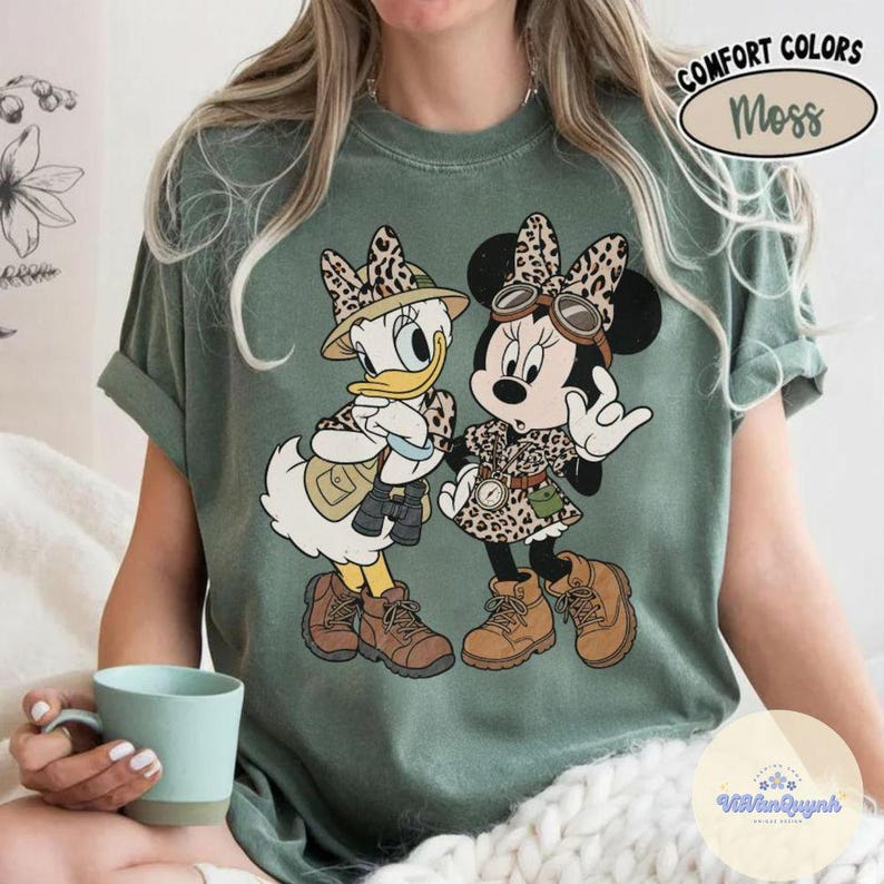 May include: A moss green t-shirt featuring a graphic of Daisy Duck and Minnie Mouse in safari outfits. The characters wear leopard print hats and clothing, with brown boots. The text "COMFORT COLORS Moss" is in the upper right corner.