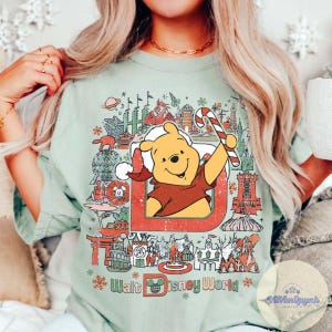 May include: Sage green t-shirt featuring a graphic of Winnie the Pooh in a Santa hat holding a candy cane, with the text "Walt Disney World" and various park icons. The design is framed by a red border and snowflakes.