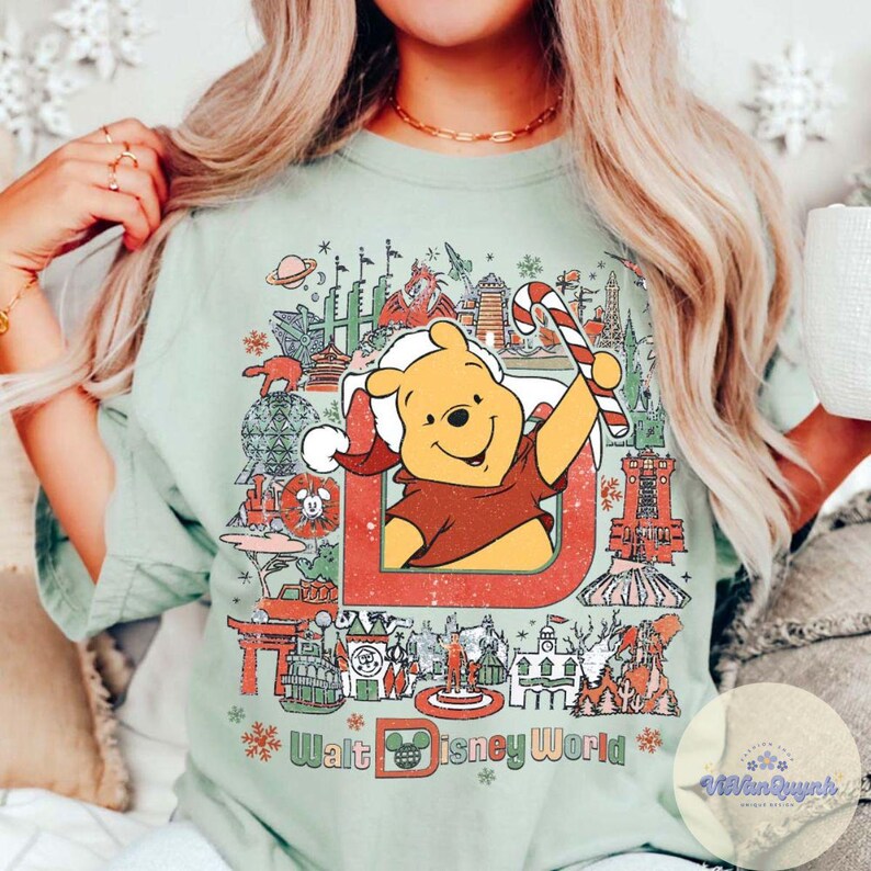 May include: Sage green t-shirt featuring a graphic of Winnie the Pooh in a Santa hat holding a candy cane, with the text "Walt Disney World" and various park icons. The design is framed by a red border and snowflakes.