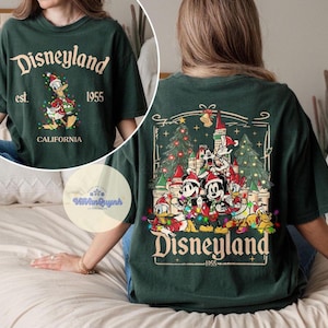 May include: Dark green t-shirt with "Disneyland" text and a graphic of Donald Duck in a Santa hat. The back features a Christmas scene with Mickey Mouse, Minnie Mouse, and other Disney characters. The text "Disneyland 1955" is also visible.