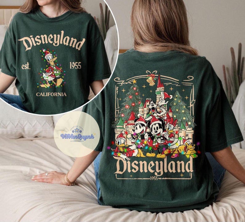 May include: Dark green t-shirt with "Disneyland" text and a graphic of Donald Duck in a Santa hat. The back features a Christmas scene with Mickey Mouse, Minnie Mouse, and other Disney characters. The text "Disneyland 1955" is also visible.