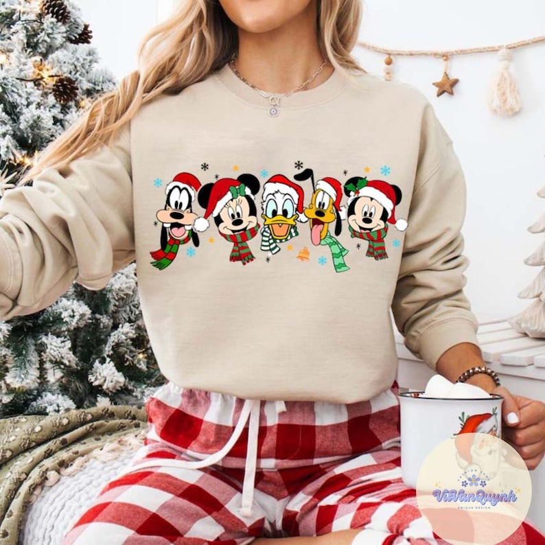 May include: Beige sweatshirt featuring a festive graphic of Disney characters, including Mickey Mouse, Donald Duck, and Pluto, all wearing Santa hats and scarves. The characters are arranged in a row, with snowflakes and stars around them. The sweatshirt is paired with red and white plaid pajama pants.