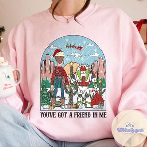 May include: Pink sweatshirt with a graphic featuring two cartoon characters in a desert scene with Christmas decorations. The text "YOU'VE GOT A FRIEND IN ME" is printed below the image. The design includes a Santa hat and a cactus.