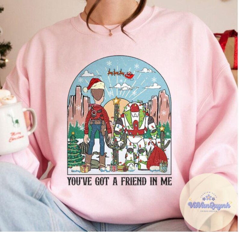 May include: Pink sweatshirt with a graphic featuring two cartoon characters in a desert scene with Christmas decorations. The text "YOU'VE GOT A FRIEND IN ME" is printed below the image. The design includes a Santa hat and a cactus.