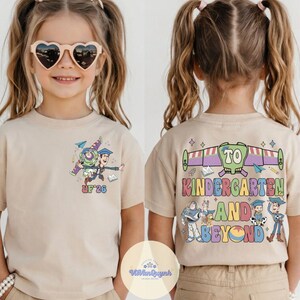 May include: A tan short-sleeved t-shirt featuring a colorful graphic of cartoon characters and the text "UP '26" on the front. The back of the shirt has the text "TO KINDERGARTEN AND BEYOND" with additional cartoon characters.