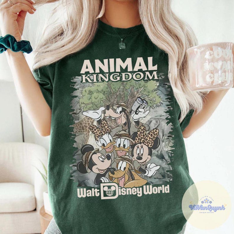K&ouml;nnte beinhalten: Dunkelgr&uuml;nes T-Shirt mit der Aufschrift "ANIMAL KINGDOM" und "Walt Disney World". Das Shirt hat eine Grafik von Mickey, Minnie, Donald, Daisy, Goofy und Pluto. Die Grafik hat ein Safari-Thema.
